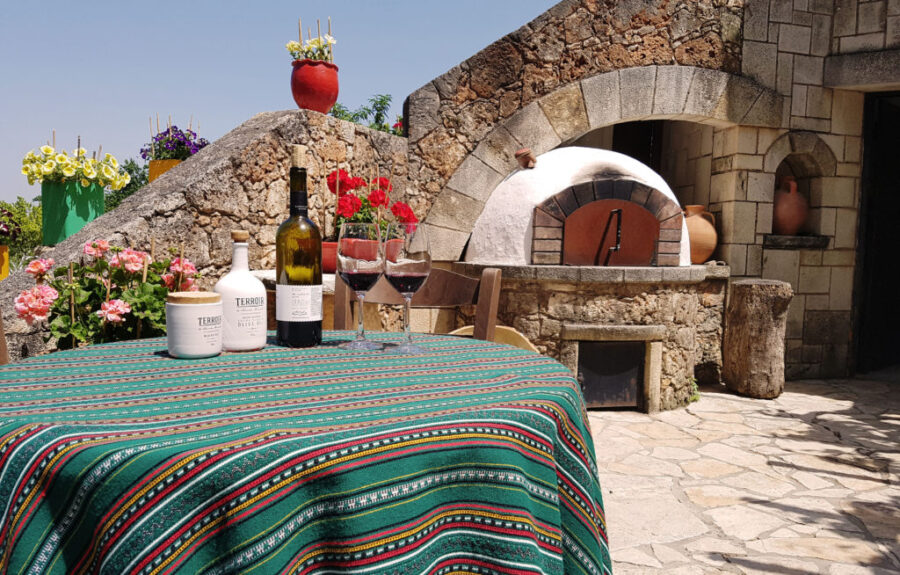 Rustic courtyard with wine, flowers, and stone oven under a sunny blue sky.