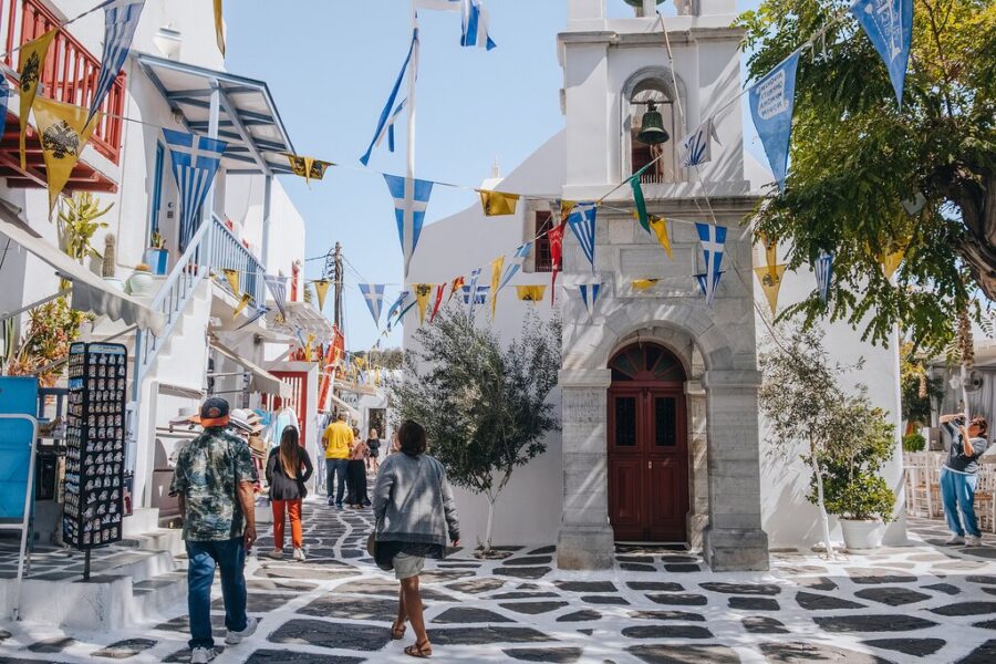 Charming Mediterranean village scene with cobblestone street, vibrant flags, and cultural landmarks.