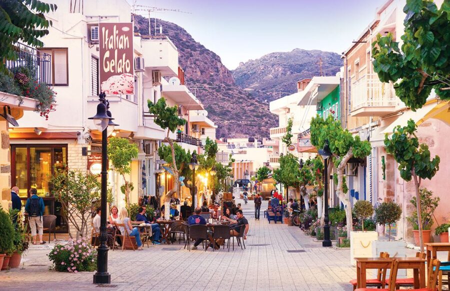 Charming Mediterranean street scene with pastel buildings, gelato shop, outdoor dining, and mountain backdrop.