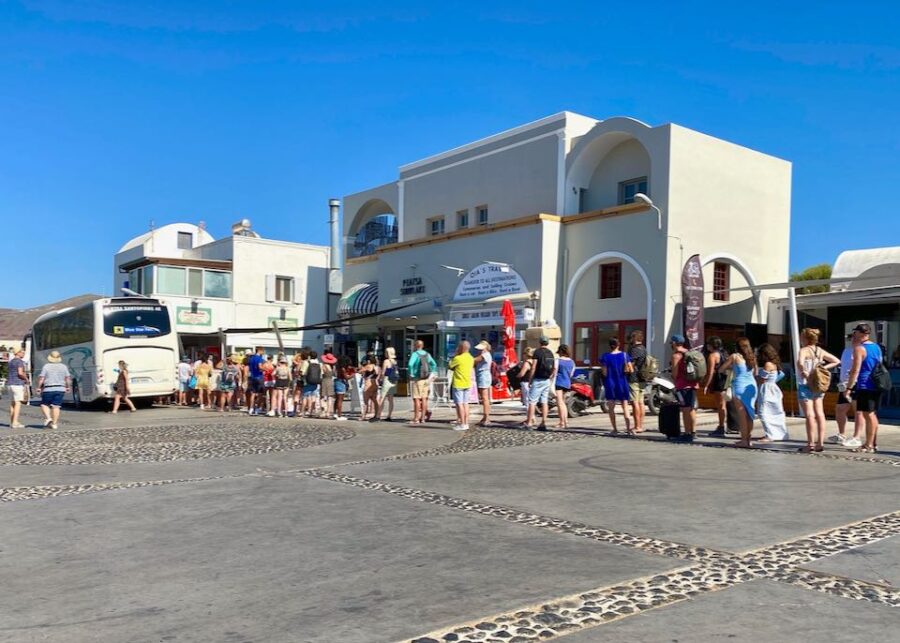 Bustling Mediterranean scene: tourists queue for bus; whitewashed buildings, red shutters in background.