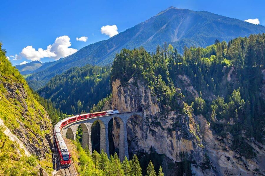 Red train crossing majestic viaduct in lush Swiss mountain landscape under clear blue skies.