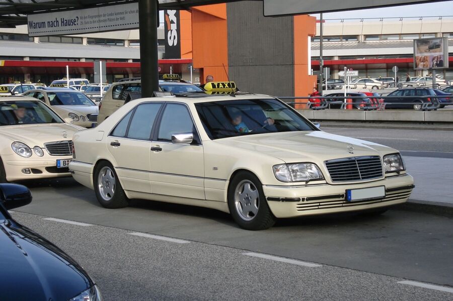 Mercedes-Benz taxi at busy airport taxi stand with overhead canopy.
