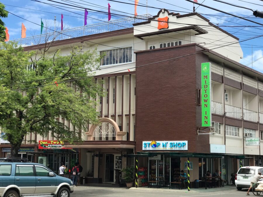 Midtown Inn street facade with storefronts, balconies, colorful flags, and parked cars.