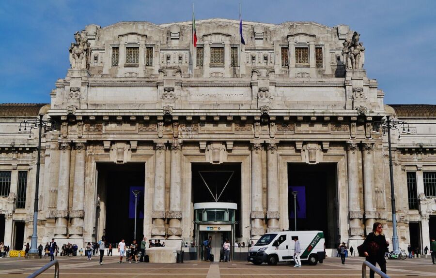 Grand Milano Centrale Station facade with statues, flags, and bustling city life.