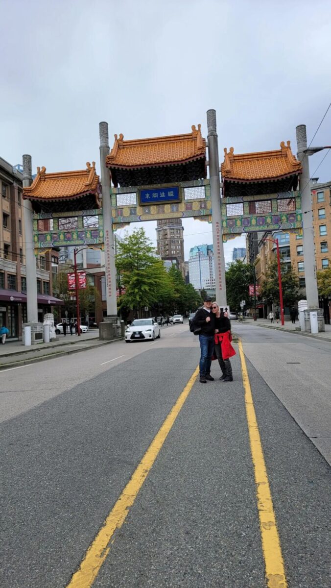 Two people are positioned in the center of the road at Millennium Gate, Vancouver, with urban surroundings visible.
