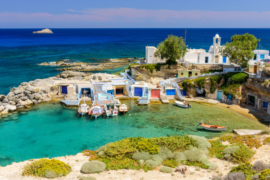 Scenic view of Milos Island's coastal village, showcasing whitewashed homes, colorful garages, and small boats in a serene bay.