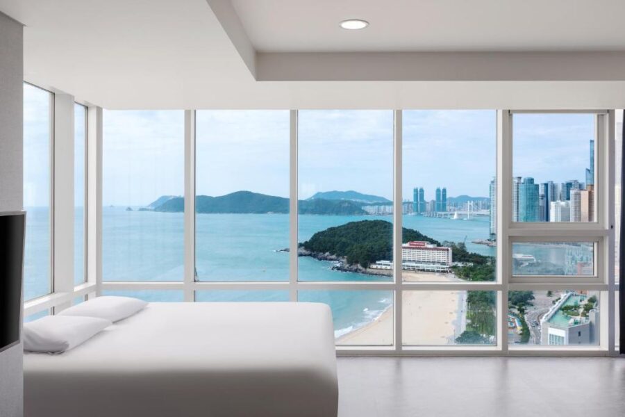 Minimalist Busan hotel bedroom with floor-to-ceiling windows, ocean view, beach, islands and city skyline.