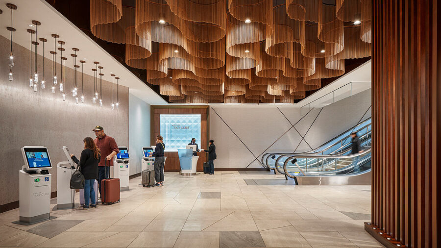 Modern airport lounge with sleek design, travelers, self-service kiosks, and wooden ceiling accents.
