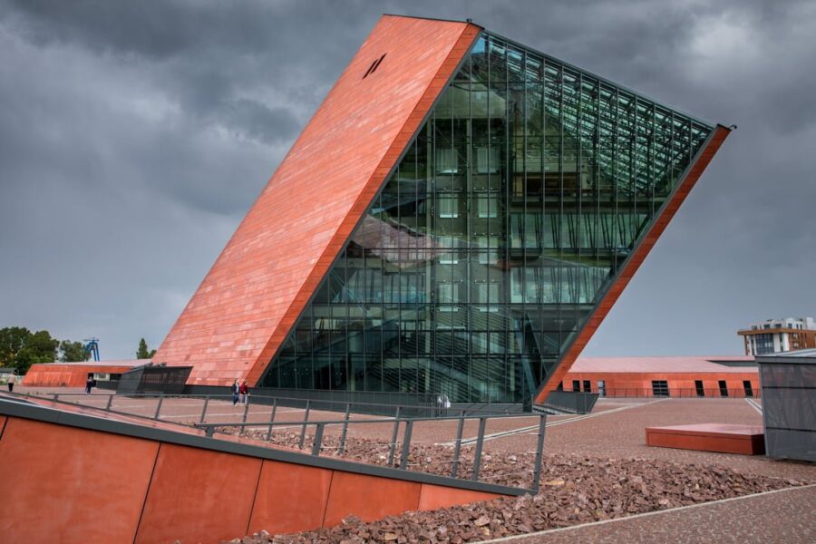 Modern angular museum with glass façade, reddish-brown exterior, landscaped surroundings, dramatic sky.