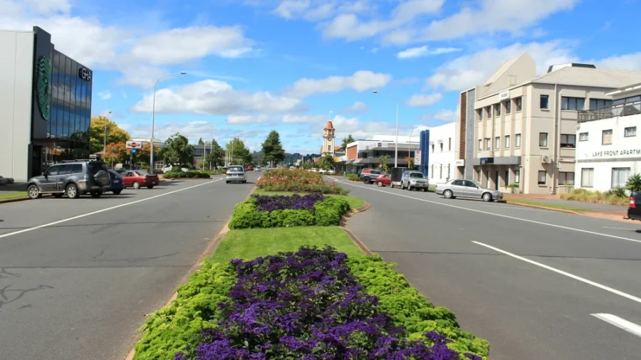 Modern Rotorua boulevard with flower-filled median, apartments, shops, streetlights, cars, and blue sky.
