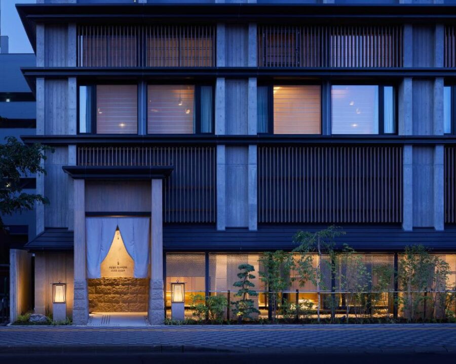 Modern Japanese-style boutique hotel facade at dusk with warm lights, entrance noren, and greenery.