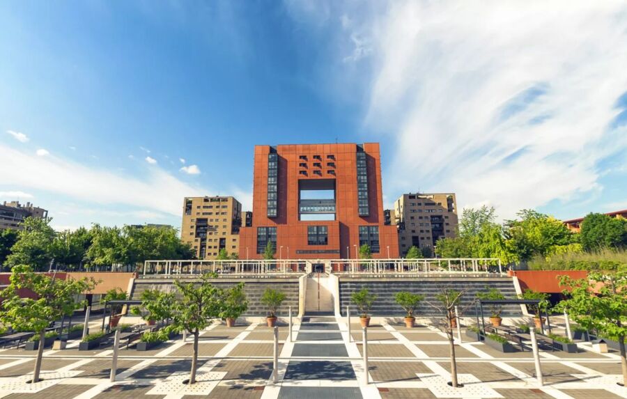 Modern building with red facade, geometric design, and expansive plaza with greenery.