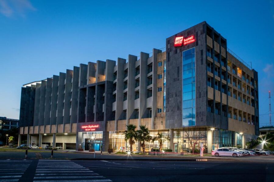 Modern city business hotel exterior at dusk with lit signage, palm trees and quiet street.