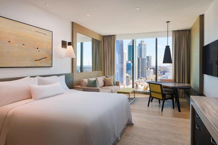 Modern luxury hotel room with king bed, city skyline view, work desk and sofa seating.