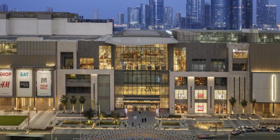 Luxury shopping mall exterior at dusk with glass façade, palm trees, and city skyscrapers.