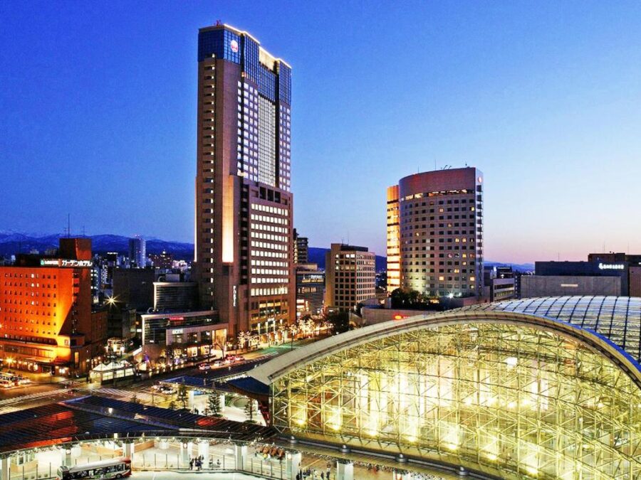 Modern city skyline at dusk with glass train station, skyscrapers, traffic and mountain backdrop.