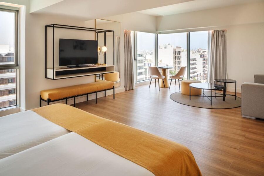 Modern hotel room with twin beds, minimalist decor, and panoramic city view from large windows.