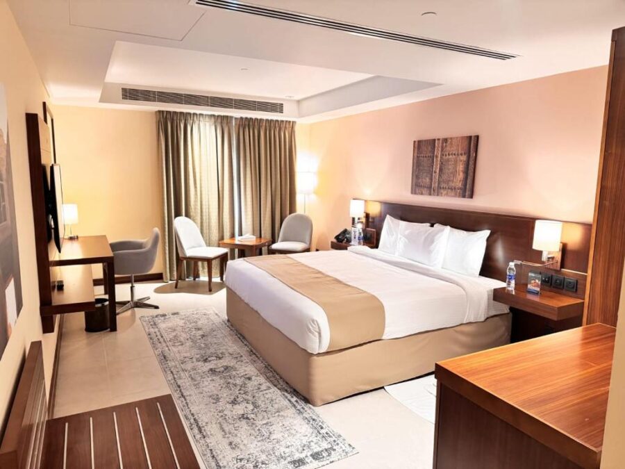Modern hotel room in Salalah with king bed, workspace, seating area, warm lighting, contemporary decor.