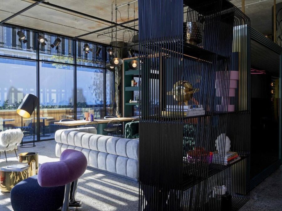 Modern industrial hotel lounge with velvet furniture, gold accents, floor-to-ceiling windows and shelving.