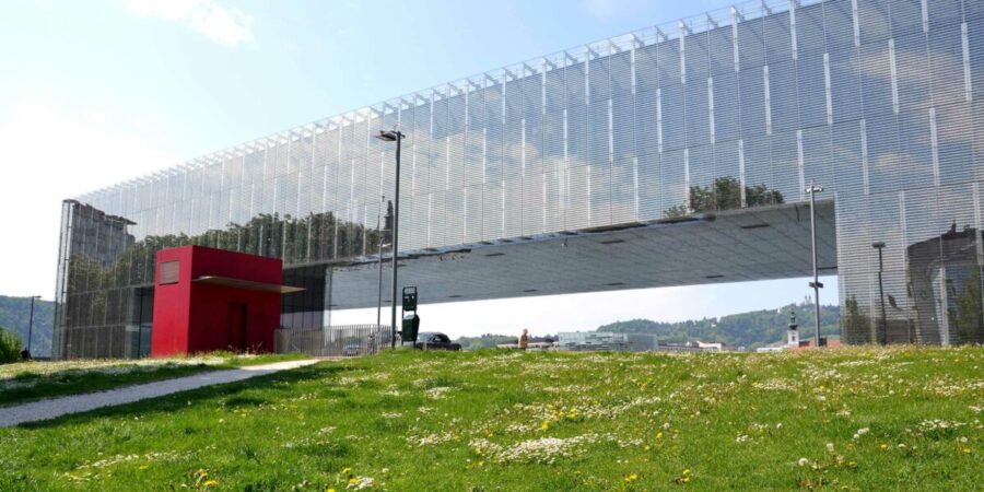 Modern cantilevered building with metal mesh facade over grassy lawn and path.