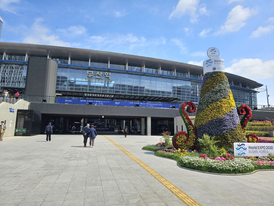 Busan Station plaza with modern glass architecture, flower display and World Expo 2030 Busan promotion.