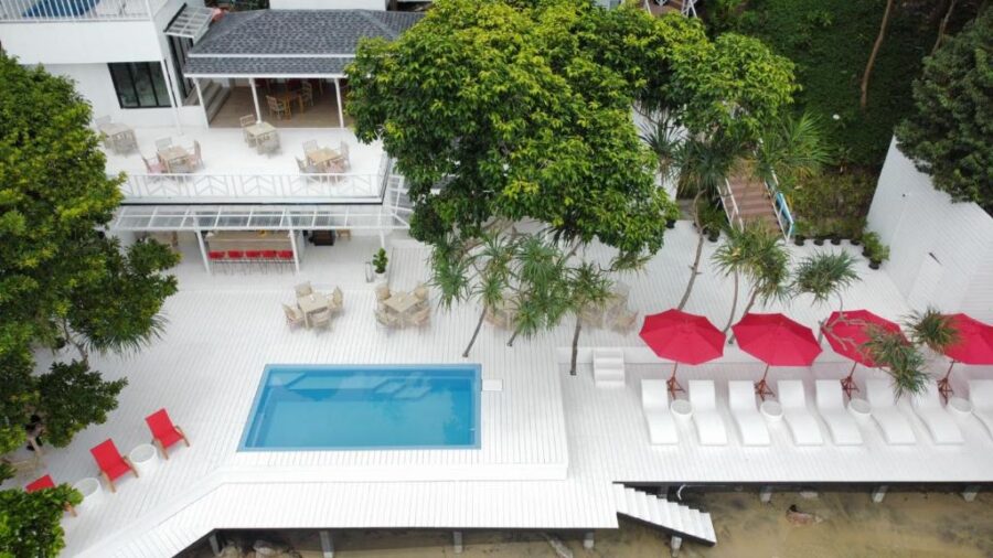 Modern seaside resort with white pool terrace, red loungers, palm trees and oceanfront luxury.