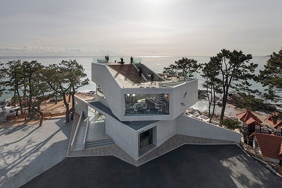 Modern concrete seaside terrace building with ocean view, rooftop deck, and coastal trees at sunset.