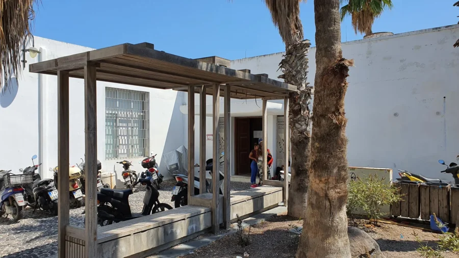 Modern building exterior with wooden shelter, motorcycles, palm tree, and foliage in warm climate.