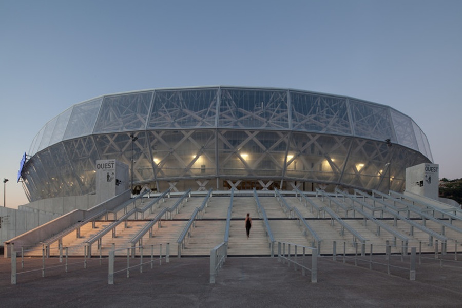 Modern stadium with transparent façade and geometric design at dusk with a lone figure.