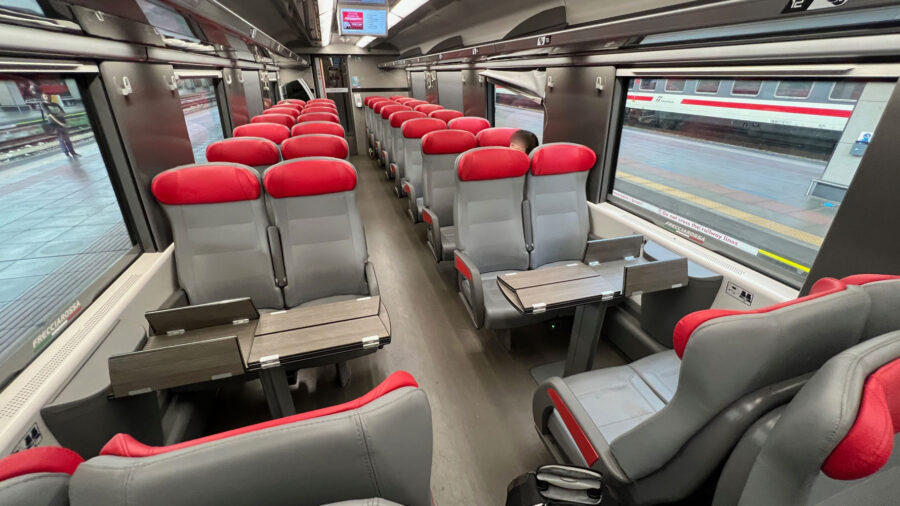 Modern train carriage interior with gray seats, red headrests, and large windows.