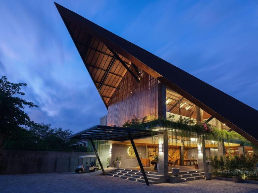 Modern tropical resort building with dramatic triangular roof, warm wood lighting, and lush greenery at dusk.