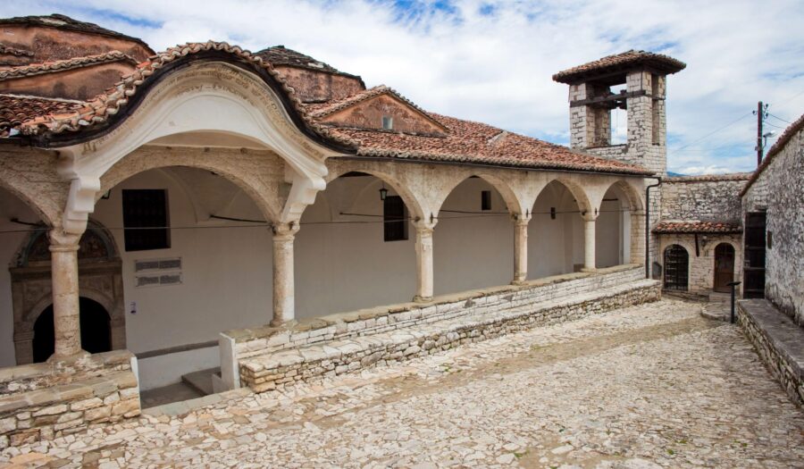 Historic monastery courtyard with arches, stone bell tower, and cobblestone path under blue sky.