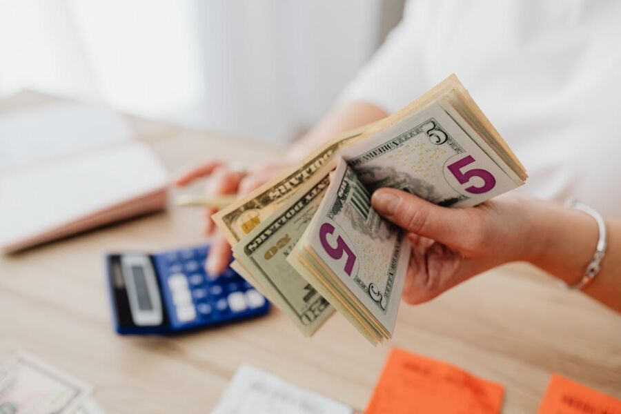 Person counting five-dollar bills with calculator, notes, and notebook for financial planning.