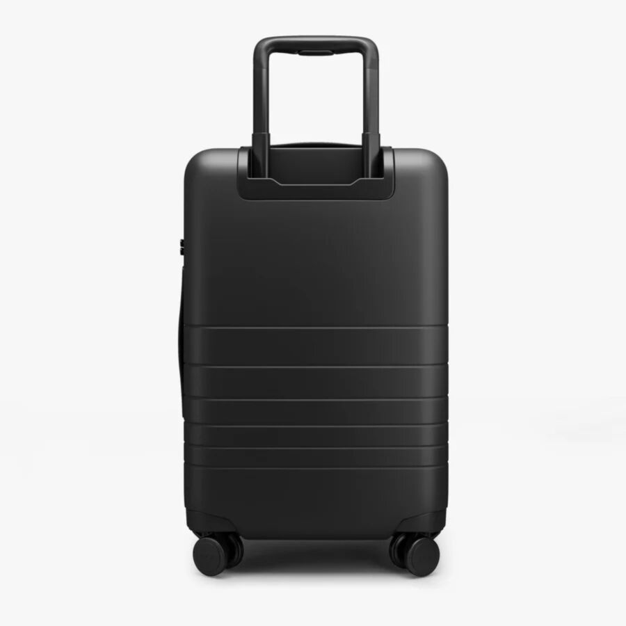 Monos Carry-On Black hard-shell suitcase on four wheels, featuring horizontal grooves and a telescopic handle. It conveys a modern, minimalist design.