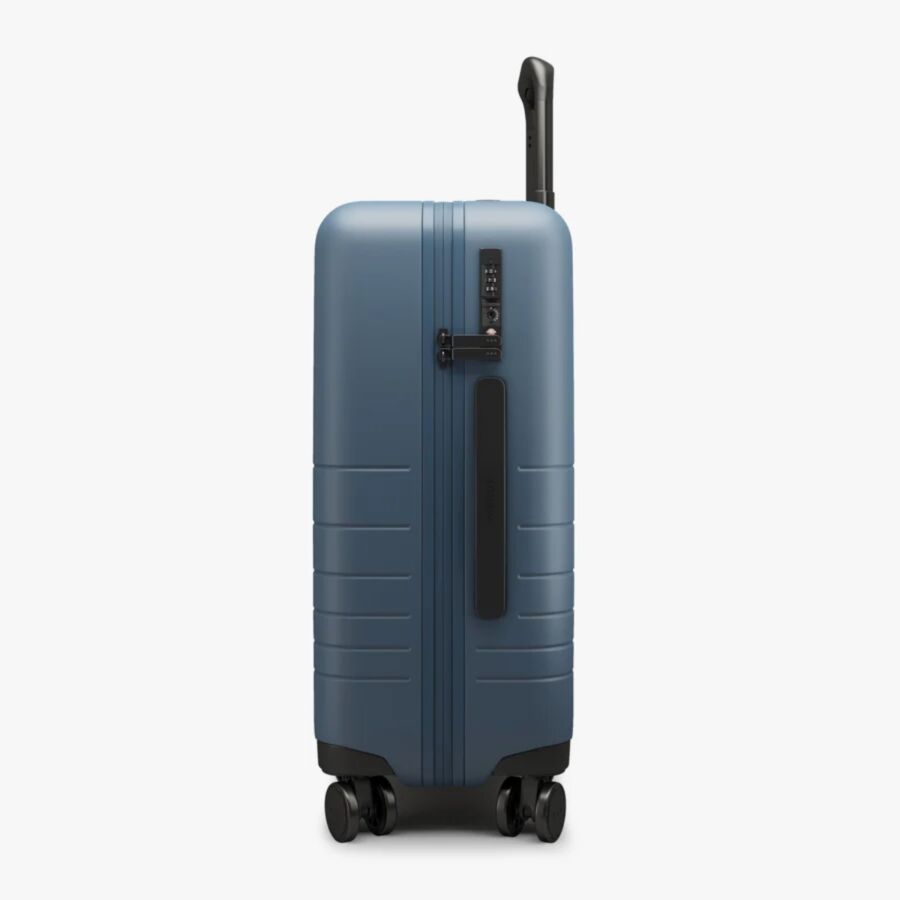 Monos Carry-On Ocean Blue hard-shell suitcase standing upright, featuring a black handle and four wheels. Side view with a visible lock against a white background.