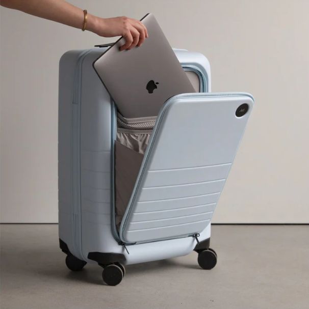 Person reveals a laptop from a blue Monos Carry-On Pro suitcase, highlighting modern travel convenience.