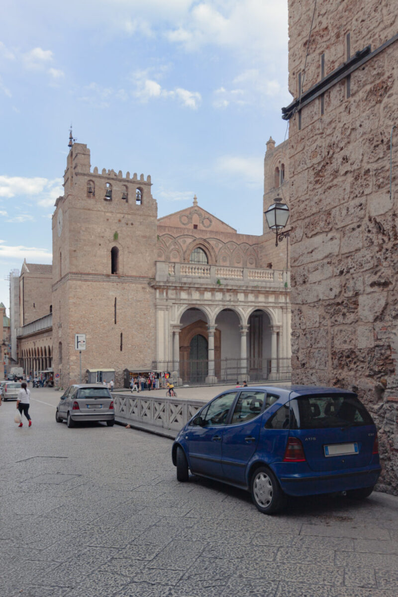 Blue car is parked on a street next to the Monreale Cathedral, surrounded by greenery.