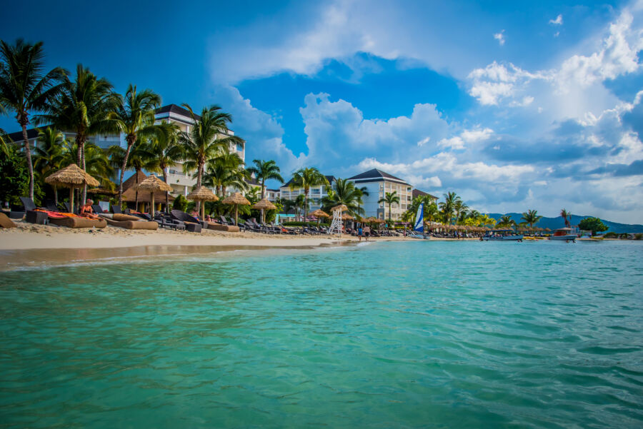 Montego Bay's beach features soft sand, clear blue water, and lush palm trees under a bright, sunny sky