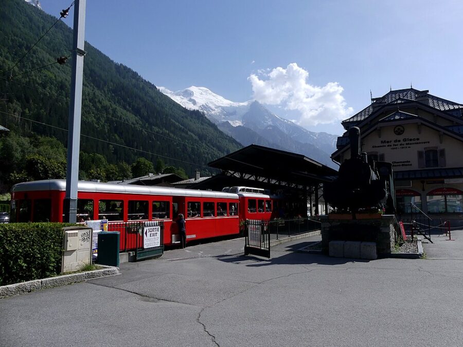 The Montenvers Railway features a red train parked at the station, with stunning mountain scenery in the background.

