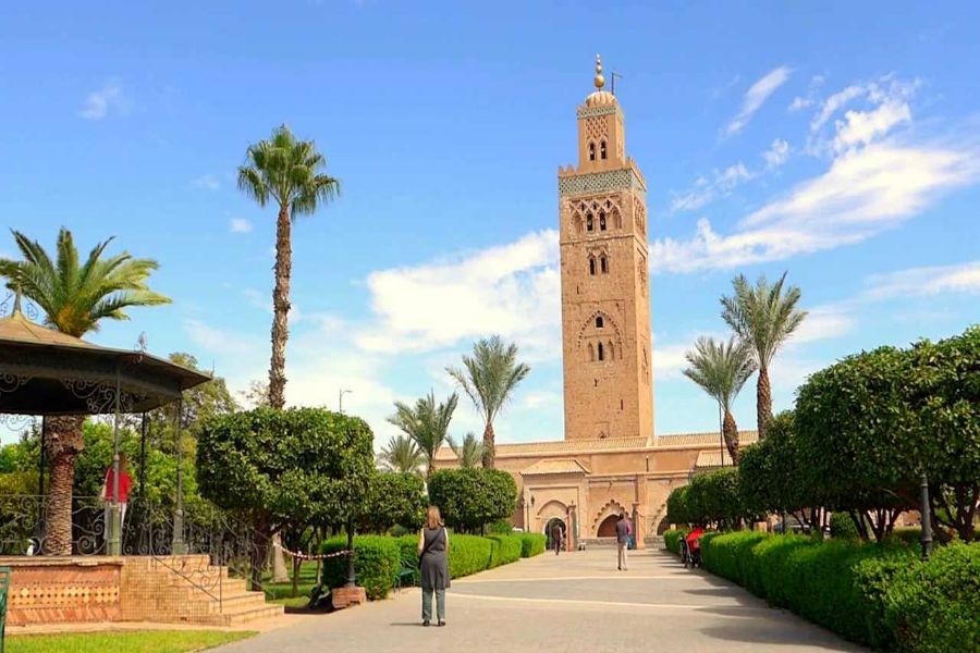 Historical Moroccan minaret with lush gardens, palm trees, blue sky, and strolling visitors.