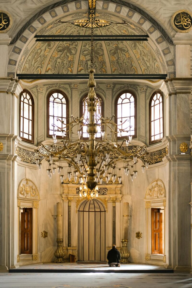 Opulent mosque interior featuring chandelier, Islamic art, and a person praying before mihrab.
