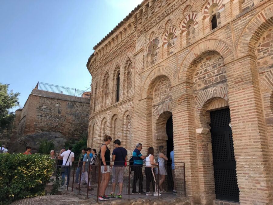 People stand in line outside a Mosque of Cristo de la Luz in Toledo historic brick building with intricate arches and patterns. The scene is lively under a clear blue sky.