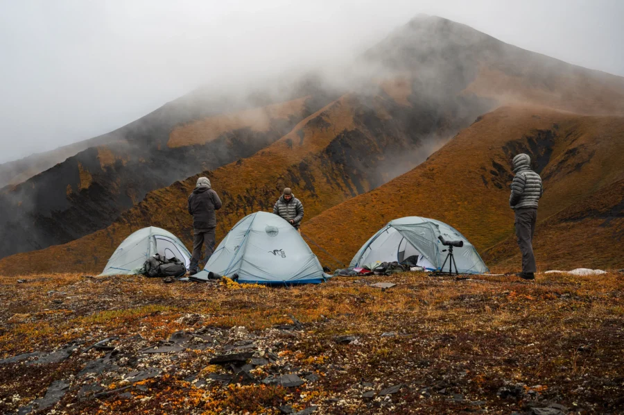 Mountain campsite with tents, rugged terrain, and misty hills in the background.