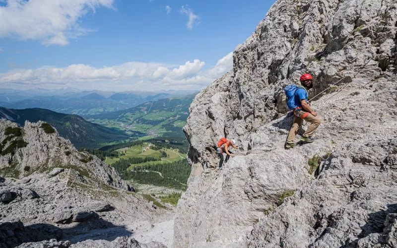 Via ferrata climbers on limestone ridge with cable above Sexten Dolomites valley.