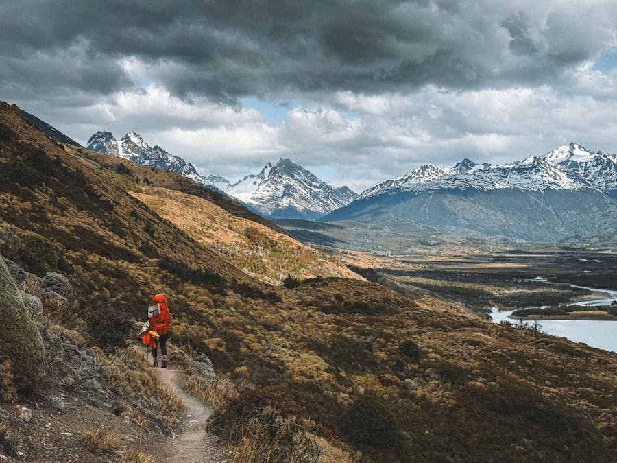 Solo hiker on rugged Patagonian mountain trail overlooking dramatic snow-capped peaks and winding river.