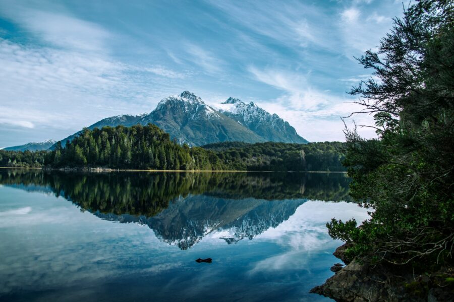 Snowy Patagonian mountain reflecting in calm lake with evergreen forest and clear blue sky.