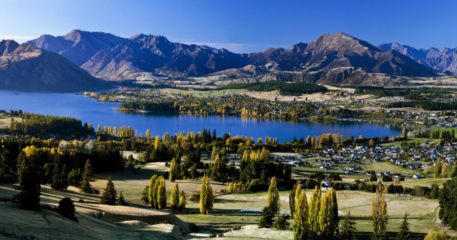 Panoramic mountain lake valley townscape with blue water, autumn trees, and distant rugged peaks.