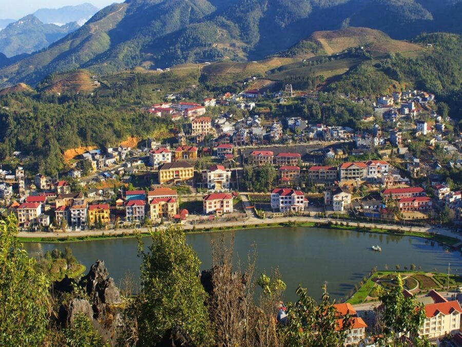 Panoramic view of Sapa mountain town with lakeside buildings, terraced hillsides, and forested peaks.