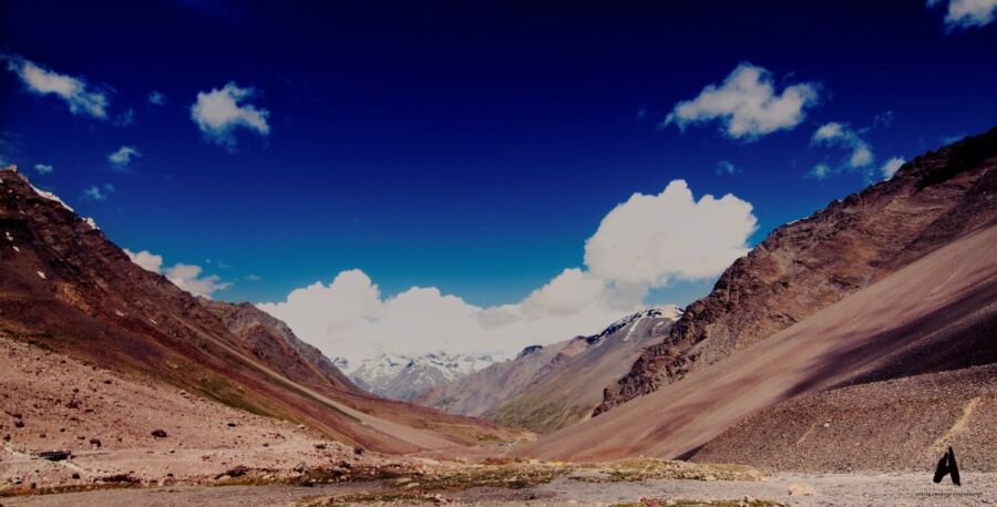 Mountainous landscape with blue sky, scattered clouds, rocky terrain, and snow-capped peaks.