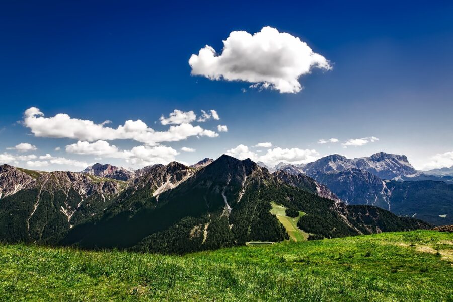 Alpine mountain landscape with green valleys, coniferous forests, and blue sky with clouds.